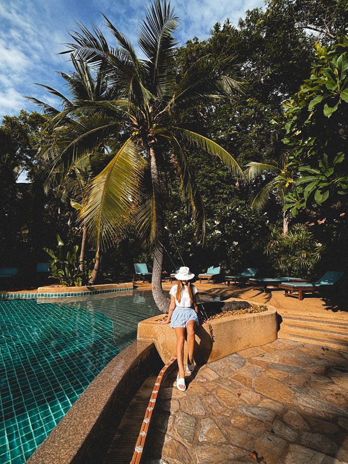 A woman in summer attire relaxes by a tropical pool surrounded by lush greenery and palm trees.